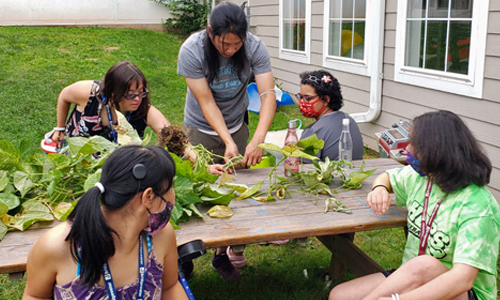 J-Cares interns and staff at a picnic table piled with leafy green plants at the Shimon and Sara Birnbaum Jewish Community Center